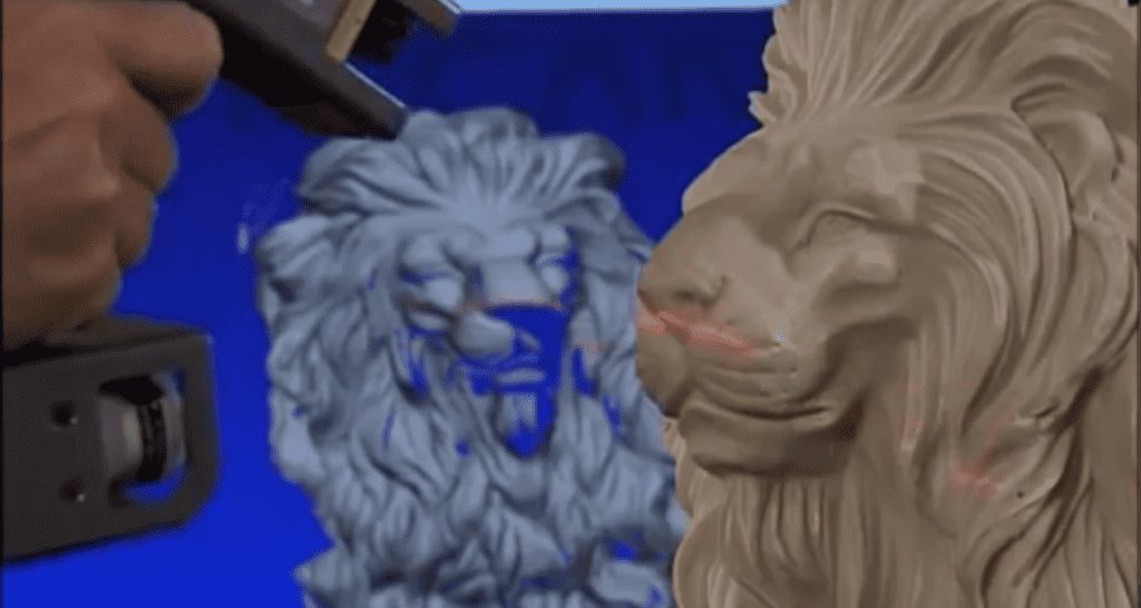 A hand holding a 3D scanning device emitting a red laser grid onto a lion sculpture to capture its detailed geometry.